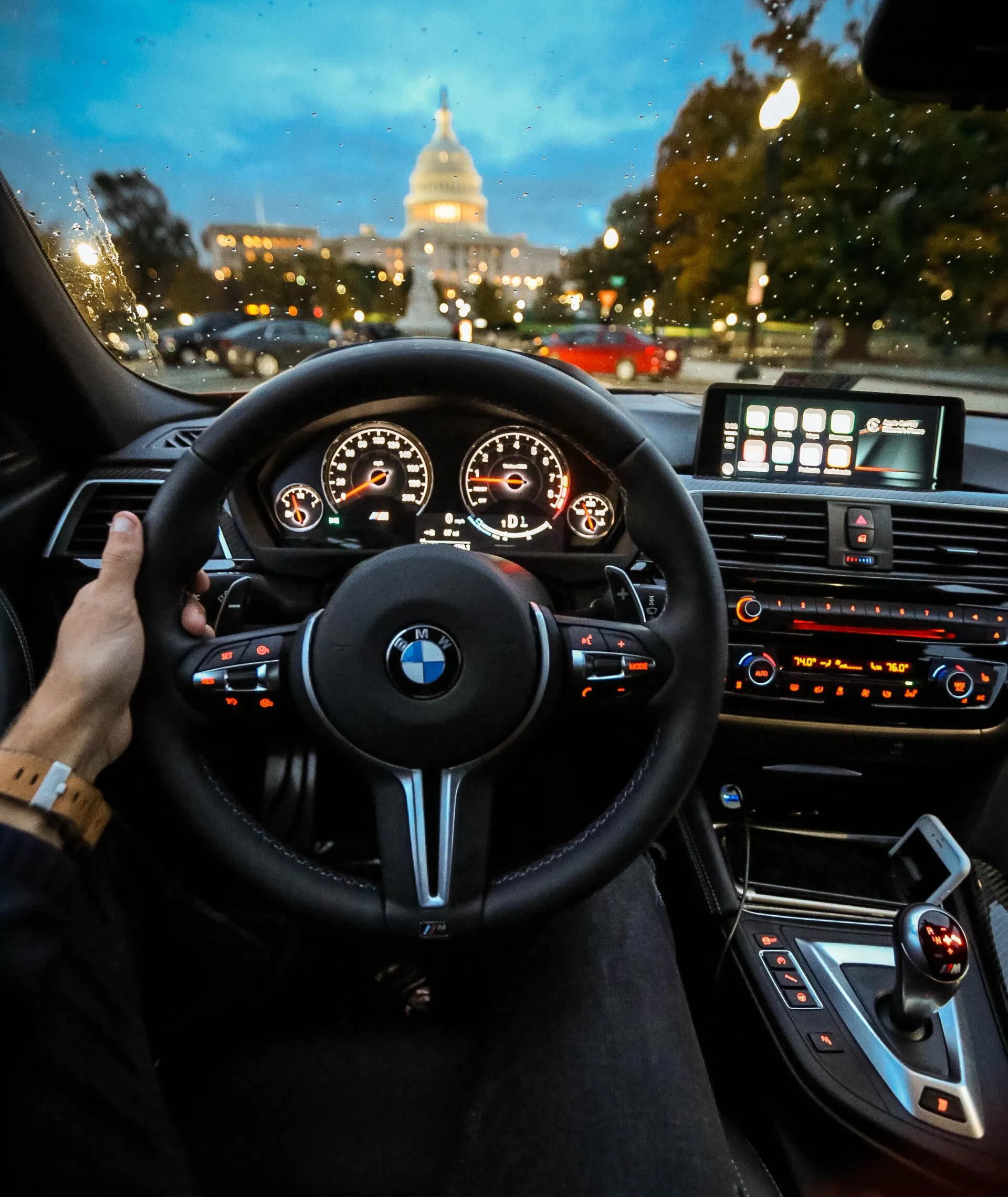 The driver's-seat view from inside a car, a hand on the steering wheel, approaching a lit domed building at dusk in the rain.