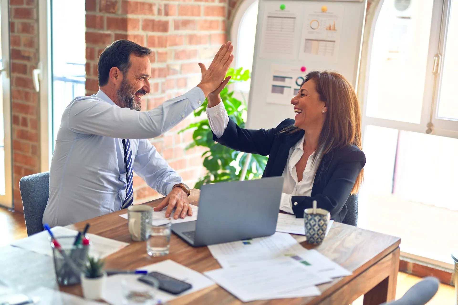 Two business professionals smiling and high-fiving over a desk with a laptop and reports.