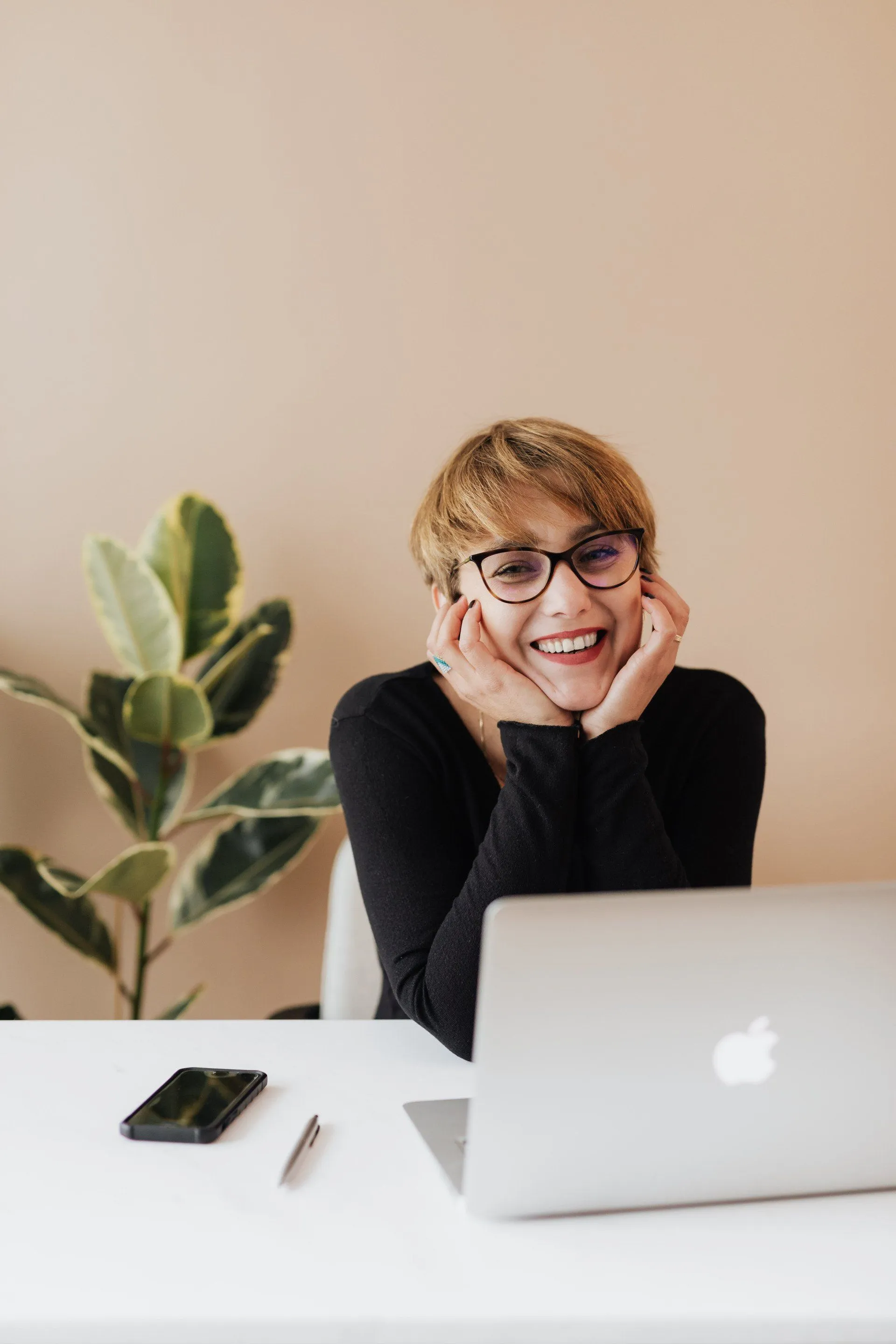 A smiling woman in glasses resting her chin on her hands at a desk with a laptop, phone, and a houseplant.