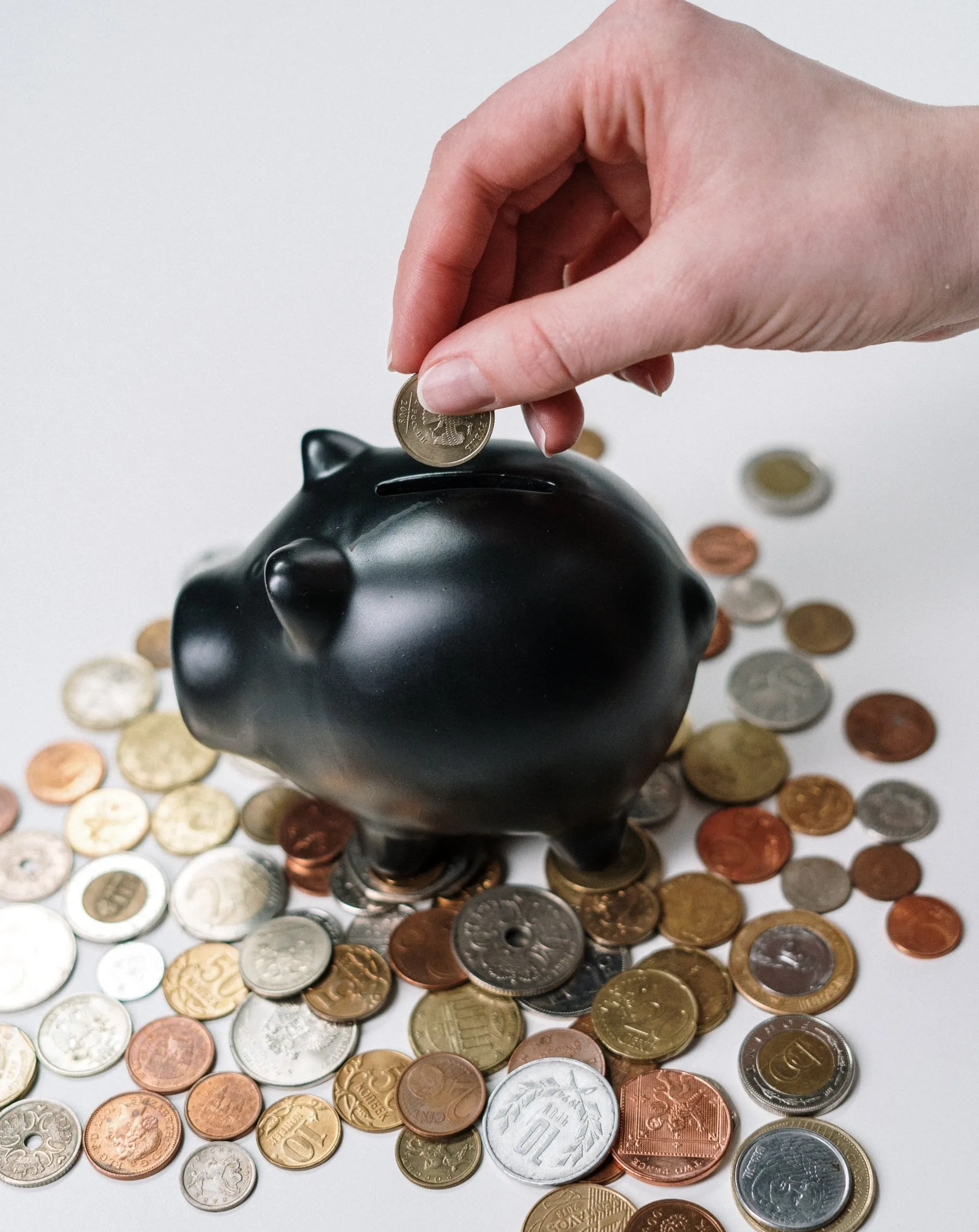 A hand dropping a coin into a black piggy bank surrounded by scattered coins.