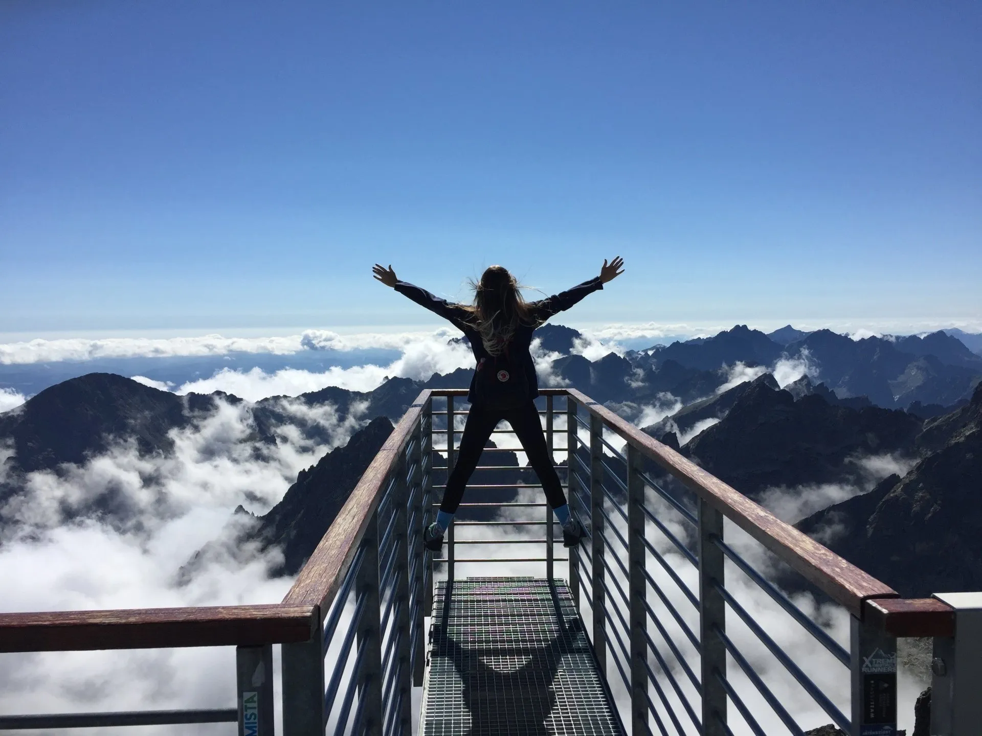 A person standing with arms outstretched on a mountaintop viewing platform above the clouds.